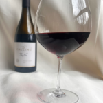Red wine poured to the widest part of the glass with bottle in the background on neutral fabric backdrop