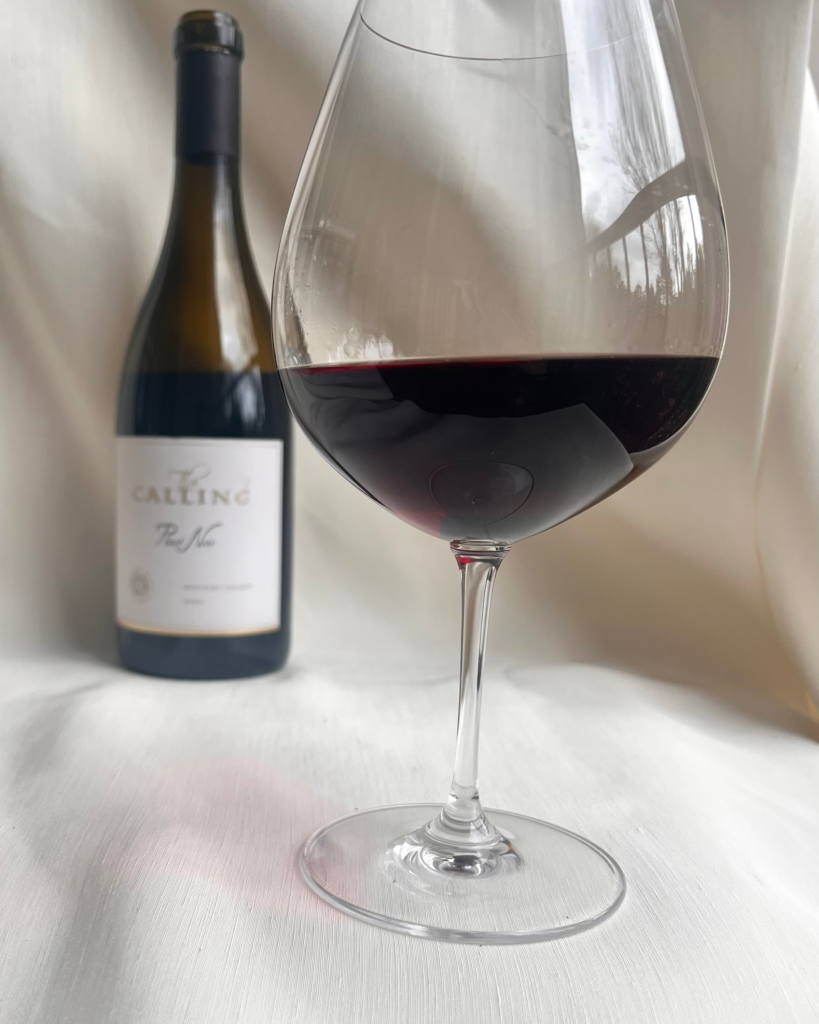 Red wine poured to the widest part of the glass with bottle in the background on neutral fabric backdrop